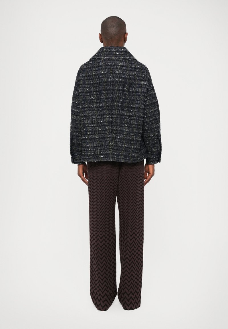 Schwarze, strukturierte Jacke mit lockerer Passform und weitem Kragen, kombiniert mit braunen, zigzag-musterierten weiten Hosen. Sichtbare Nähte und Akzente.