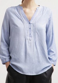 Kobieta ubrana w jasnoniebieską bluzkę z długim rękawem, z plisowanymi detalami i trzema guzikami przy dekolcie w kształcie V, z rękami schowanymi w kieszeniach czarnych spodni.