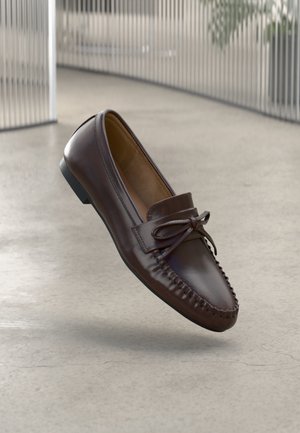 Brune lærloafers med avrundet tupp, sløyfedetalj og dekorative sømmer langs vampen. Flat svart gummisåle.