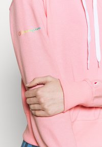 Rosa hoodie i mjukt tyg med ribbade ärmslut. Har en vit dragsko och ett broderat "Champion"-logotyp på ärmen.