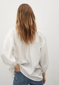 White, textured blouse with gathered back and balloon sleeves, paired with blue jeans. Features a relaxed fit and subtle striped pattern.