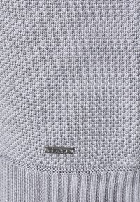 Gros plan sur un tissu tricoté gris clair avec une petite étiquette métallique rectangulaire indiquant le nom de la marque cousue près du bord inférieur.