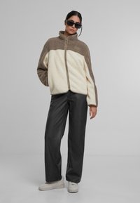 Chaqueta de forro polar con cremallera en color crema y marrón, que presenta un cuello alto, un acabado texturizado y bolsillos laterales, combinada con pantalones oscuros y zapatillas blancas.