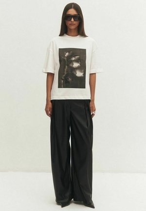 Kobieta w oversize białej koszulce z czarno-białym abstrakcyjnym kwiatowym nadrukiem, czarne szerokie spodnie, duże czarne okulary przeciwsłoneczne i pierścionki.
