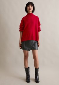 Roter gestrickter Rollkragenpullover mit gerippten Details, kombiniert mit einem kurzen schwarzen Leder-Wickelrock und kniehohen schwarzen Stiefeln.