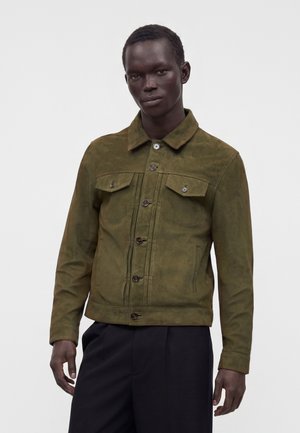 Man draagt een olijfgroene jas met knopen en klepzakken en een zwarte broek, staand tegen een effen lichte achtergrond.