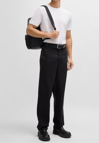 Biała koszulka z krótkim rękawem, czarne spodnie, czarny pasek i czarne obuwie. Niesie czarny plecak, stoi na jasnym tle.