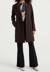 Mörkbrun kappa i ullblandning, med två knappar och sidofickor, kombinerad med en mönstrad blus och svarta utsvängda byxor.