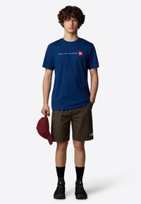 Camiseta de algodón azul marino con texto y logotipo blancos, combinada con pantalones cortos marrones. Gorro rojo sostenido en la mano, calcetines negros y zapatos negros completan el atuendo.