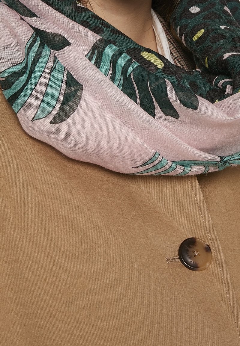 Écharpe à motifs rose clair et vert foncé enroulée autour du cou sur un manteau couleur chameau avec un bouton rond visible.