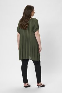 Tunique couleur vert olive à manches courtes retroussées, texture légère et design fluide, portée avec un pantalon noir ajusté et des chaussures à petit talon.