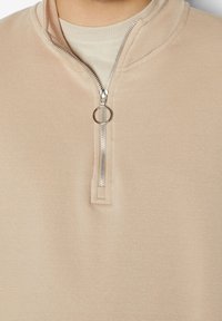 Suéter beige con diseño de media cremallera, que cuenta con una cremallera plateada y un tirador en forma de anillo. Textura suave con un cuello de canalé por debajo.