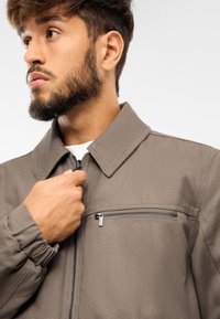 Šedá bunda na zips s golierom, prednou zipsovou kapsou a zberanými rukávmi. Hladká látka s matným povrchom. Model drží zips.