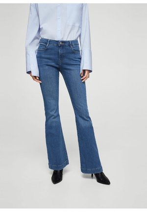 Femme portant un jean évasé bleu, une chemise bleu clair boutonnée avec de longs poignets, et des bottes noires à talons pointus, debout devant un fond uni.