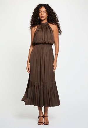 Femme aux cheveux bouclés portant une robe midi marron sans manches à col halter et des talons marron à brides, debout devant un fond uni.