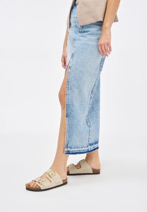 Gonna maxi in denim di colore blu chiaro, con uno spacco laterale e orlo sfrangiato. Sandali beige con due fibbie e suola simile al sughero.