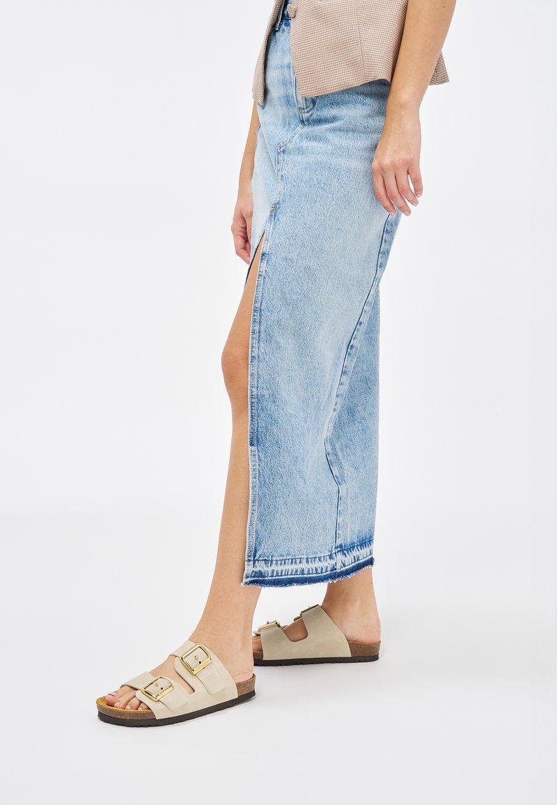 Gonna maxi in denim di colore blu chiaro, con uno spacco laterale e orlo sfrangiato. Sandali beige con due fibbie e suola simile al sughero.