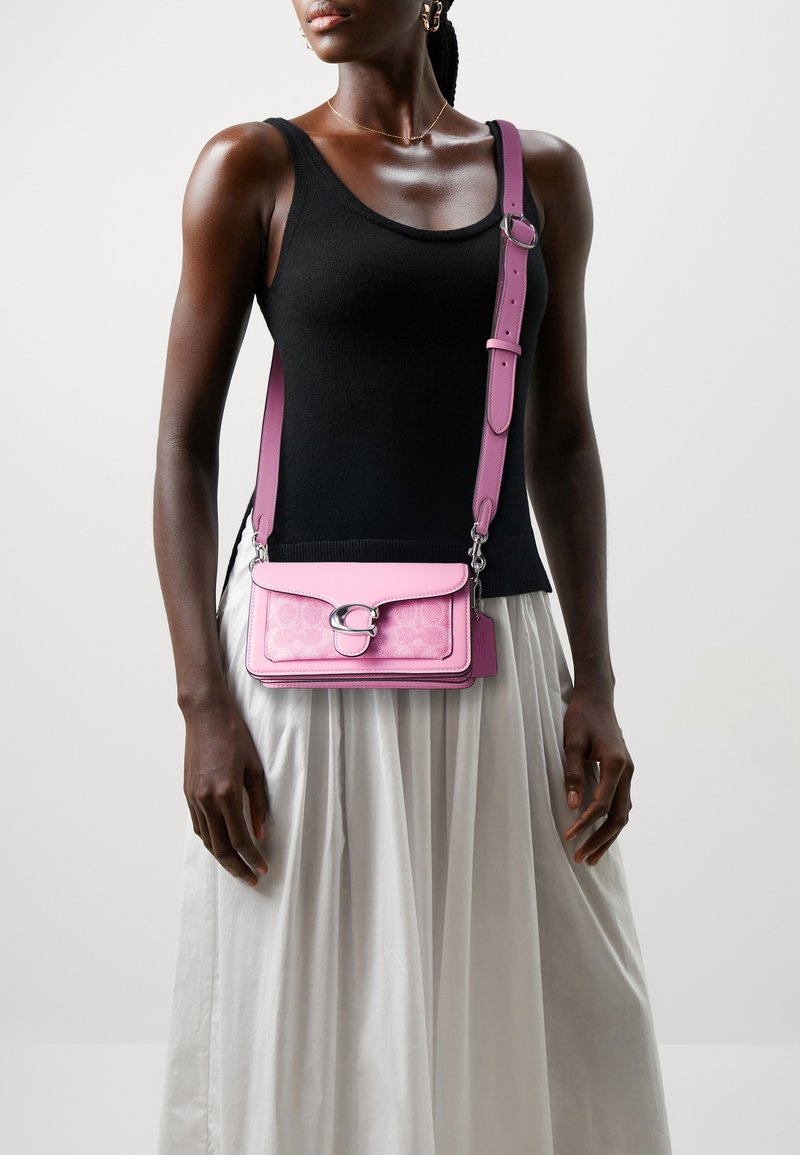Femme portant un haut noir sans manches et une jupe blanche, avec un petit sac bandoulière rose à large bandoulière réglable.
