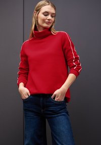 Sudadera roja con alto cuello y rayas blancas a los lados, combinada con vaqueros de denim azules. Tejido suave, diseño casual, ajuste relajado.