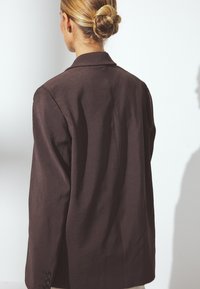 Brązowa dopasowana marynarka o gładkiej teksturze, z wyciętymi klapami i zapinanymi mankietami. Charakteryzuje się czystą, dobrze skrojoną sylwetką i minimalnymi detalami.
