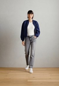 Marineblå bomberjakke over en hvit T-skjorte, parret med lysegrå rettløpende jeans og hvite sneakers. Tregulv, nøytral bakgrunn.