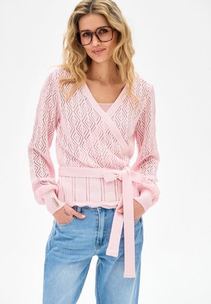 Kobieta o blond, falowanych włosach, nosząca duże okulary, różowy zapinany sweter w wzory z paskiem oraz jasnoniebieskie jeansy, z rękami w kieszeniach, lekko się uśmiechająca.