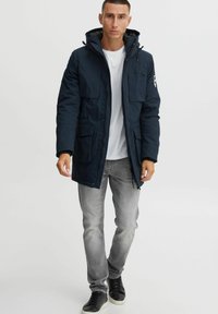 Marineblaue Parka mit Reißverschluss, mehreren Taschen, verstellbarem Kapuze und grauem Innenfutter. Kombiniert mit hellgrauen Jeans und schwarzen Sneakers.