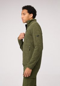 Giacca in pile verde scuro con colletto alto, tasca sul petto con zip e logo ricamato sulla manica; superficie a maglia texturizzata.