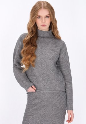 Young woman with long wavy hair wearing a gray ribbed knit turtleneck sweater and matching patterned knit skirt against a white background.