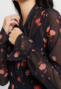 Blusa in chiffon nero con motivi floreali rosa, caratterizzata da un collo con fiocco, maniche a sbuffo con polsini elasticizzati e una texture trasparente.