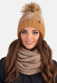 Gebreide mosterdkleurige beanie met een pompon en crème accenten, gecombineerd met een gestructureerde beige sjaal. Beiden stralen een gezellige en warme uitstraling uit.