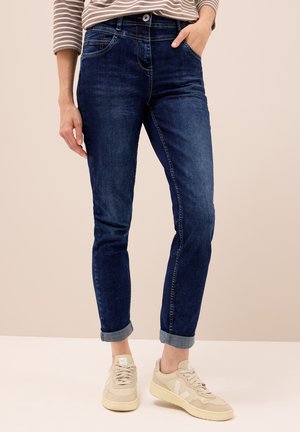 Donkerblauwe slim-fit jeans met opgerolde boorden en voorzakken, gecombineerd met lichtgekleurde sneakers met een gestructureerd ontwerp.