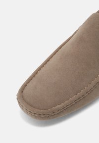 Bout avant de mocassin en daim taupe doux avec surpiqûres visibles le long du bord, présenté sur un fond clair uni.
