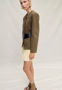 Olive green fitted blazer with a black belt, layered over a cream pleated skirt. Model wearing beige socks and black sandals. Neutral background.
