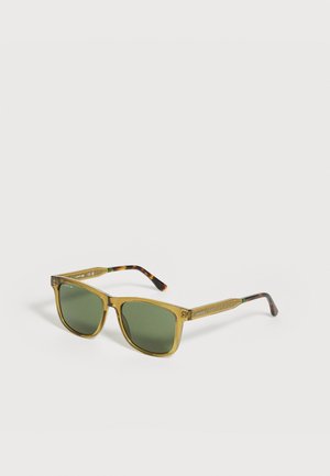 Lunettes de soleil avec verres verts, montures carrées jaunes translucides et branches à motif écaille, posées sur une surface claire.