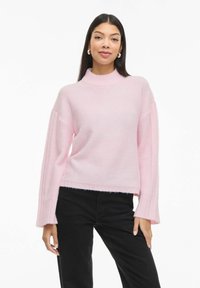 Jasnoróżowy dzianinowy sweter z wysokim dekoltem i długimi, prążkowanymi rękawami. Krócony fason z wykończeniem chwostami na dole.