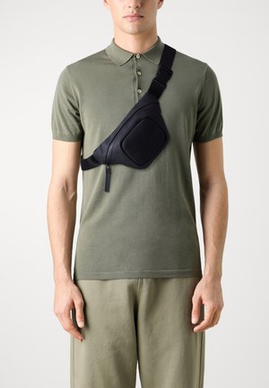 Borsa a tracolla nera in materiale liscio, con forma arrotondata, chiusura con zip e tracolla regolabile, indossata sopra una polo verde.