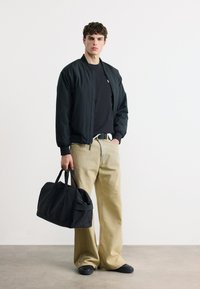 Νεαρός άνδρας που φοράει μαύρο μπουφάν τύπου μπόμπερ και μπεζ παντελόνι φαρδιάς γραμμής, κρατώντας μια μεγάλη μαύρη τσάντα duffel μπροστά σε ένα απλό λευκό τοίχο.