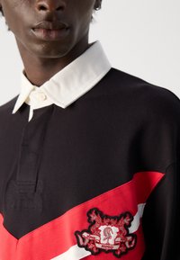 Primo piano di una persona che indossa una polo nera con colletto bianco e una striscia diagonale rossa e bianca con un ricamo a forma di scudo Hugo.
