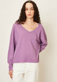 T-shirt ample à manches longues en tissu violet clair. Il présente un décolleté en V et des poignets côtelés. L'ourlet est légèrement effiloché, associé à un pantalon blanc.