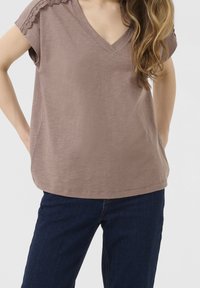 Braunfarbendes V-Ausschnitt-T-Shirt mit Spitzenverzierungen an den Schultern, aus weichem Stoff gefertigt. Getragen mit einer dunklen Jeans mit entspannter Passform und abgerundetem Saum.