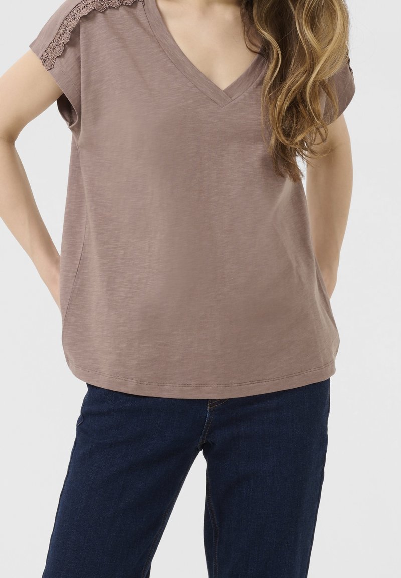 Braunfarbendes V-Ausschnitt-T-Shirt mit Spitzenverzierungen an den Schultern, aus weichem Stoff gefertigt. Getragen mit einer dunklen Jeans mit entspannter Passform und abgerundetem Saum.