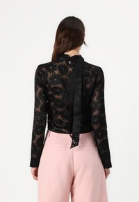 Black floral lace top with long sleeves, sheer texture, and a tied neck detail, paired with light pink high-waisted trousers.