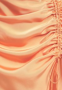 Tissu en satin pêche avec une texture lisse, présentant des détails froncés sur un côté et un cordon de serrage pour un ajustement ajustable.