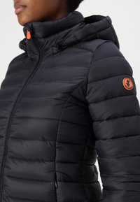 Černá péřová bunda na zip s kapucí, s horizontálním prošíváním a texturovanou látkou. Oranžový akcent na rameni.