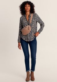 Lässiges Outfit mit einer gemusterten dunkelblauen Bluse, dunklen Jeans und braunen Stiefeletten. Eine beige Umhängetasche wird quer über den Körper getragen.