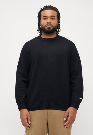Mann mit geflochtenem Haar, der einen schwarzen Pullover und beigefarbene Hosen trägt, steht vor einem schlichten weißen Hintergrund und blickt nach vorne.