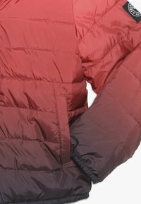 Czerwono-burgundowa kurtka puchowa z pikowanymi sekcjami, elastycznym mankietem i czarną naszywką z logo na ramieniu. Teksturowana powierzchnia.