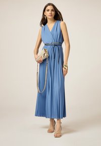 Blauwe geplooide jurk met een diepe V-hals, cinched in de taille met een decoratieve riem. Het model houdt een lichtgekleurde tas vast en draagt hakken met bandjes.