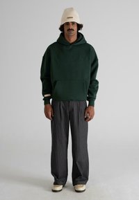 Hombre con una sudadera verde oscuro, pantalones negros a rayas, gorro crema y zapatos crema, de pie contra un fondo claro y liso.